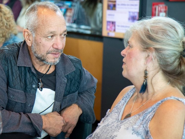 Image of An older man and woman sit closely together indoors, engaged in a serious conversation. The man wears a dark jacket and necklace, while the woman has fair hair and wears a patterned sleeveless top and earrings.