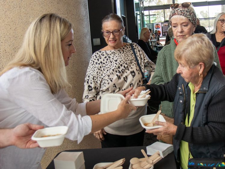 Image of A woman hands a food container to an elderly woman in a queue of people at a distribution table, with others waiting and watching in a bright indoor setting. Disposable bowls and wooden cutlery are on the table.
