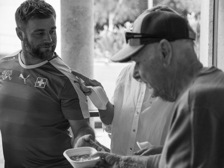 Image of A man in a Swiss football shirt hands a bowl of food to an elderly man wearing a cap, while another person holds a bottle behind them. The scene appears to be outdoors and in black and white.