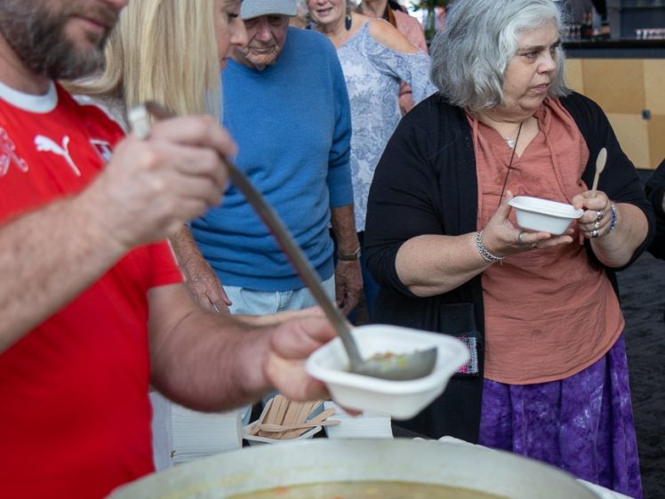Image of A man ladles soup into bowls for people queueing at an indoor event. Several adults wait their turn, holding empty bowls, while one woman receives her serving and steps away.
