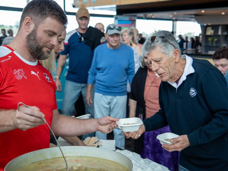 Image of A man in a red shirt ladles soup into a bowl held by an older man in a navy shirt, whilst several people wait in queue behind them in a busy indoor setting.