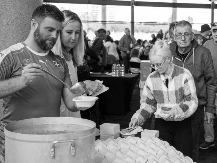 Image of A man ladles soup into a bowl for a woman, whilst another woman picks up bowls from a table stacked with containers. Several people wait in queue in the background at what appears to be a community food event.