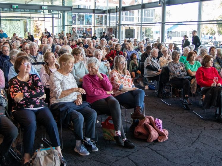 Image of A large group of elderly people sitting closely together in rows of chairs inside a bright, modern event space, attentively watching something out of shot.