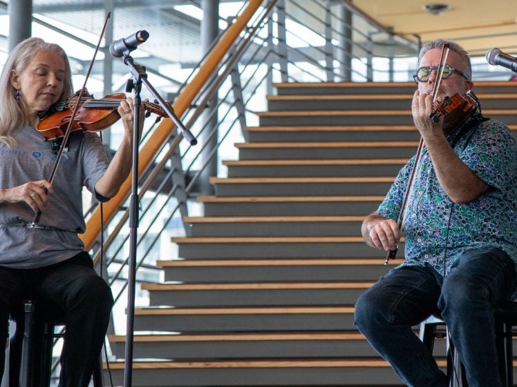 Image of Two people play violins on a stage in front of a staircase. The woman on the left stands while playing and the man on the right is seated, both focussed on their instruments. Microphones are set up in front of them.