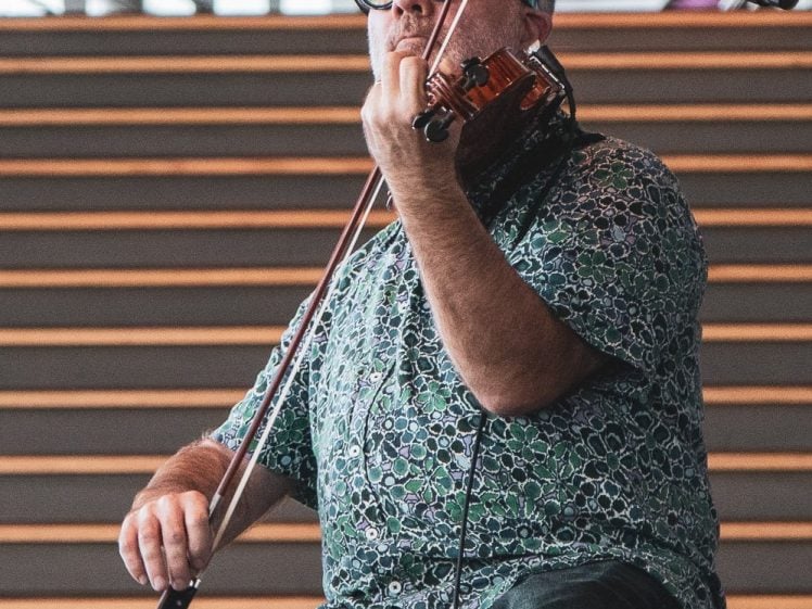 Image of A man wearing glasses and a patterned shirt plays the violin with focus, sitting in front of wide wooden steps and a microphone on a stand.
