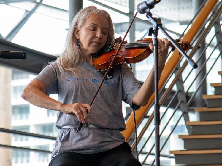 Image of A woman with long grey hair plays the violin whilst seated on a stage, wearing a grey T-shirt. A microphone is positioned in front of her, and a staircase and large windows are visible in the background.