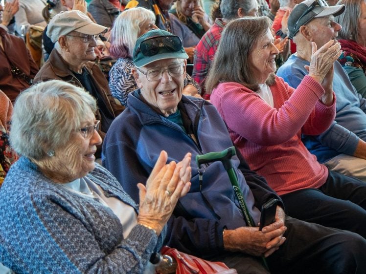 Image of A group of older adults sit closely together in an audience, smiling and clapping enthusiastically. The atmosphere appears cheerful and lively, with many people wearing casual, colourful clothing and hats.