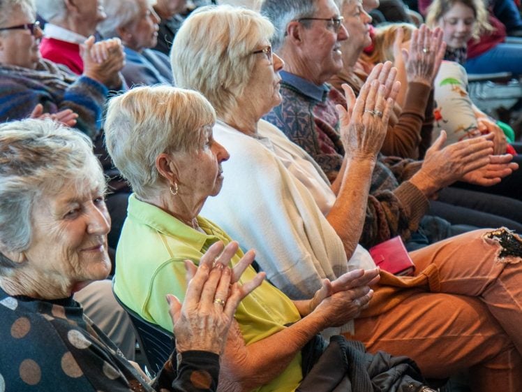 Image of A group of older adults sit closely together in rows, smiling and applauding as they watch an event or performance. The atmosphere appears cheerful and lively.