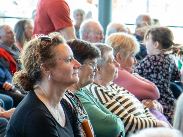 Image of A group of people of varying ages sit closely together, attentively watching something off-camera indoors. One person stands in the background, whilst most others are seated and focused forward.