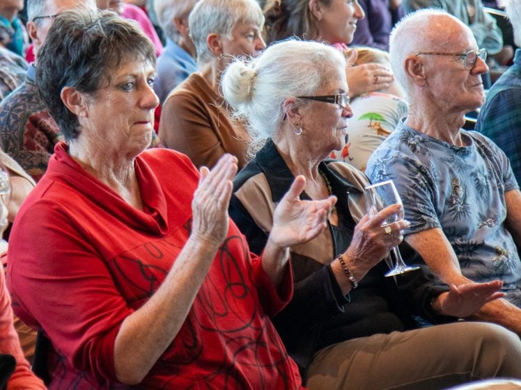 Image of A group of older adults sit closely together in an audience. A woman in a red shirt applauds, while another woman holding a glass and a man in a blue patterned shirt watch attentively.
