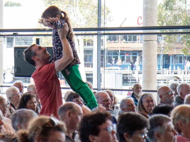 Image of A man in a red shirt lifts a smiling young girl above his head in a crowded indoor gathering, with people seated around them and large windows showing buildings outside.