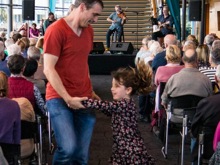 Image of A man and a young girl joyfully dance in an aisle during a live music performance, with an audience seated behind them and musicians playing on a stage in a bright, modern venue.