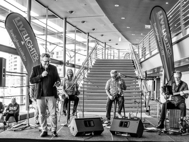 Image of A black-and-white photo of a band performing indoors in front of a staircase. Four musicians play instruments whilst one sings. Two large Livelighter banners stand on each side, with seated audience members in the background.