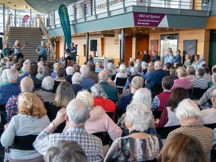 Image of A large audience of mostly older adults sits facing a stage area indoors, where musicians and singers are performing. The venue is modern with glass walls and an upstairs balcony.