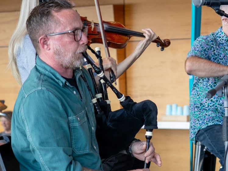 Image of A man in a green shirt plays the bagpipes while a woman behind him plays the violin. They are seated indoors, performing together on a stage with microphones nearby.