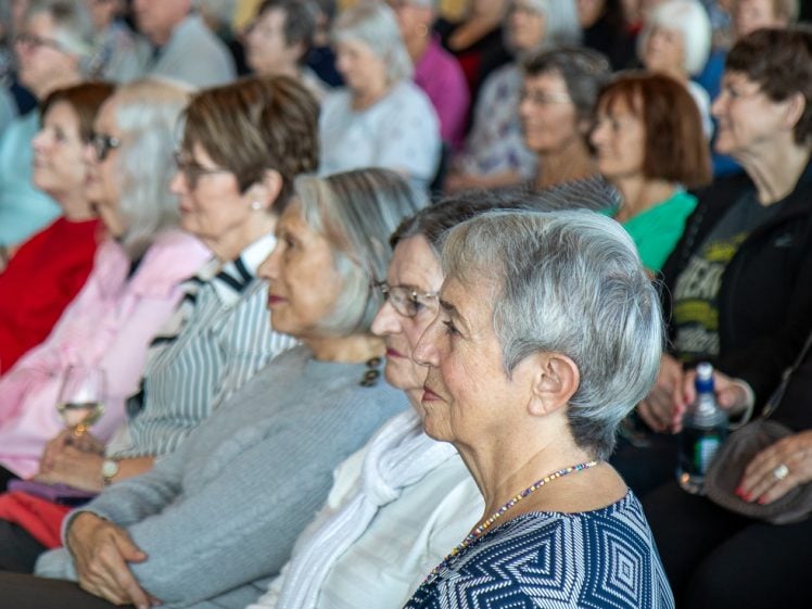 Image of A group of older adults, mostly women, sit closely together in rows, attentively watching something out of view. Most wear glasses and casual clothing, with some holding drinks. The atmosphere is focused and engaged.