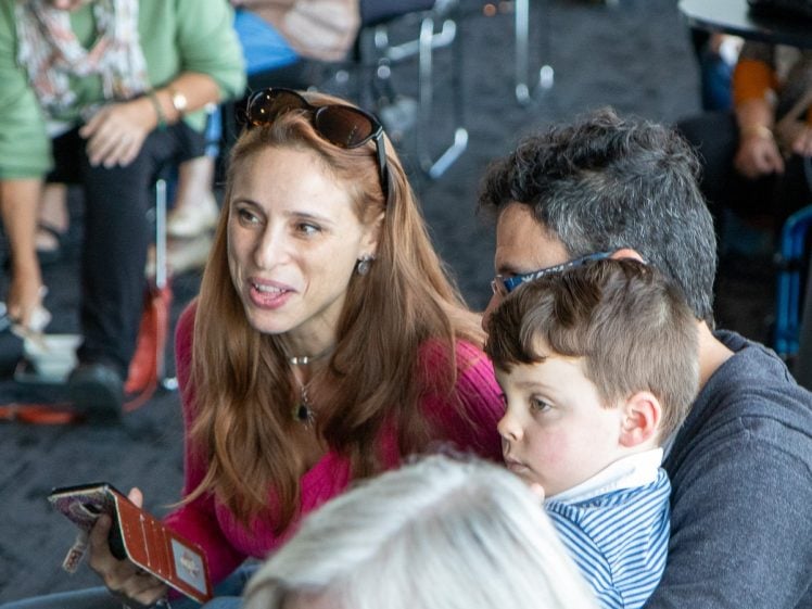 Image of A woman holding a passport smiles whilst sitting next to a man and a young boy on his lap in a busy indoor setting with other people in the background.