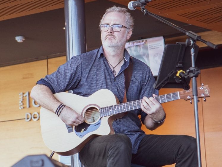 Image of A man with short grey hair and glasses sits on stage playing an acoustic guitar. He wears a dark shirt and trousers, with a microphone and music stand in front of him. The background features wood panelling and signage.