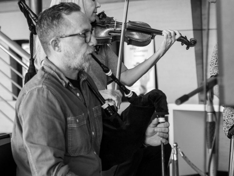 Image of A man plays the bagpipes while a woman beside him plays the violin. Both appear focused on their music in what looks like a modern indoor setting. The image is in black and white.
