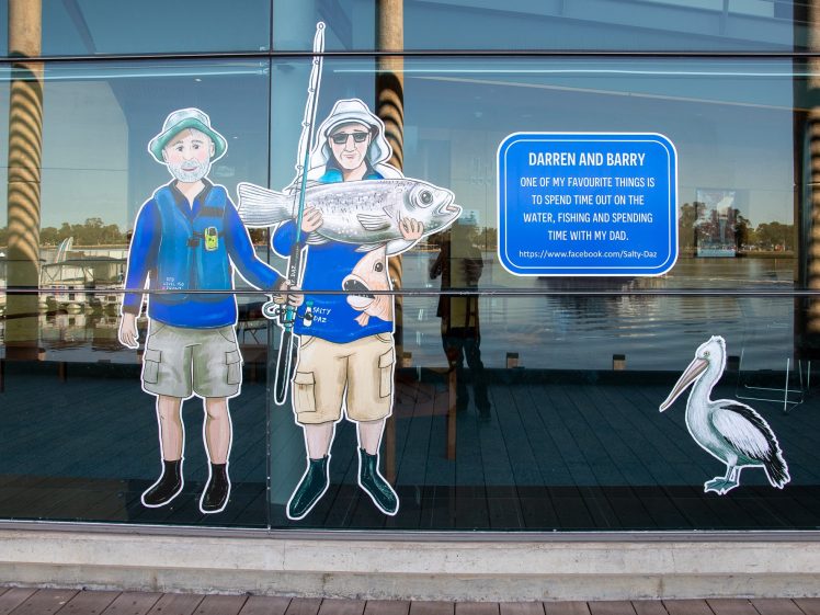Image of Two cartoon cut-outs of men with fishing gear and a large fish are displayed on a glass window. A pelican cut-out sits nearby. A blue sign shares a quote about fishing with Dad. Reflections of a waterfront can be seen through the glass.