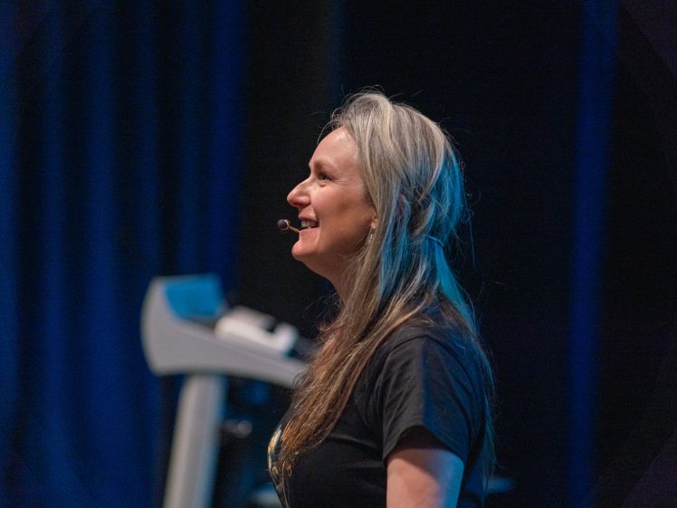 Image of A woman with long grey hair, wearing a black t-shirt and a headset microphone, smiles whilst speaking on stage with a dark background and some equipment visible beside her.
