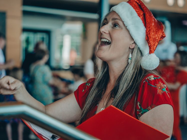 Image of A woman wearing a Santa hat and red outfit smiles and sings whilst holding a red folder, with people and festive decorations blurred in the background.