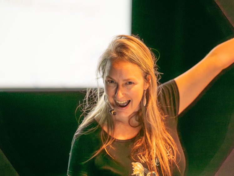Image of A woman with long blonde hair, wearing a black shirt and a headset microphone, smiles energetically with one arm raised against a blurred background.