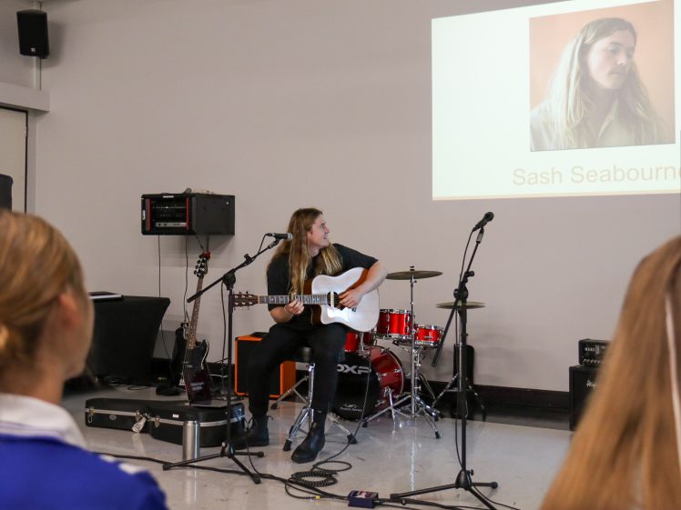 Image of A musician sits on a chair playing an electric guitar onstage, with drums and sound equipment behind them. An audience watches, and a projected slide shows a photo of the musician and the name Sash Seabourn.