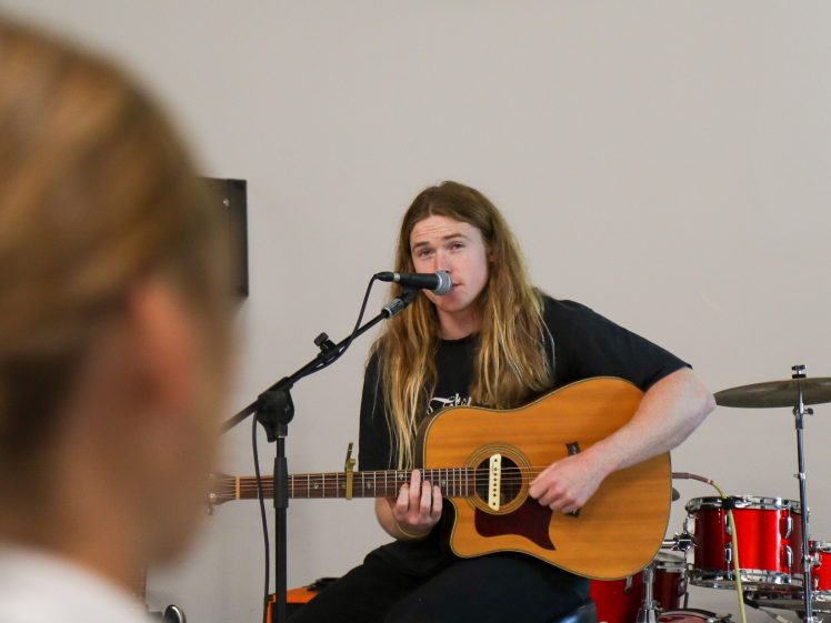 Image of A musician with long blonde hair plays an acoustic guitar and sings into a microphone on stage; a drum kit is visible in the background, and a blurred audience member is in the foreground.