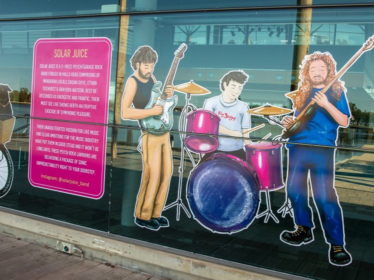 Image of A colourful mural on glass panels shows three cartoon musicians: a guitarist, a drummer, and a bassist. Next to them is a pink sign titled Solar Juice describing their solar-powered music. The reflection of the street is visible.