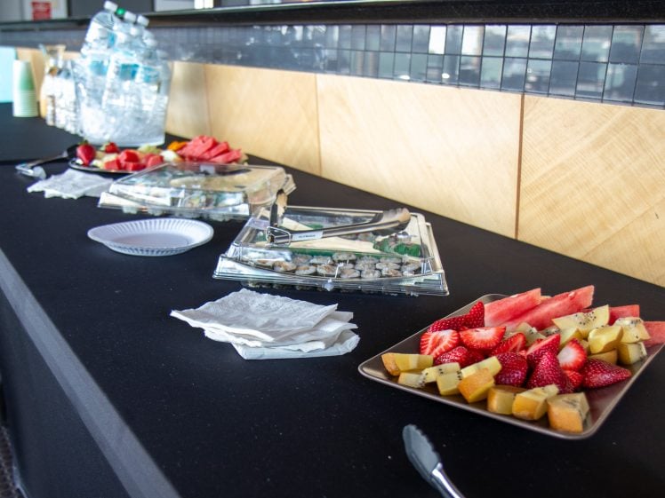 Image of A black tablecloth-covered table displays trays of cut fruit, sushi rolls, bottled water, paper plates, and serviettes in a brightly lit indoor setting.