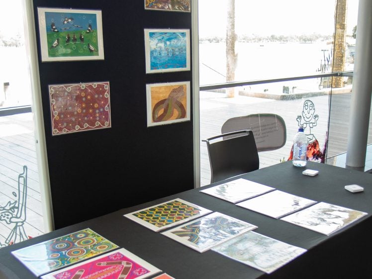 Image of A display table with colourful artwork prints arranged on top and several pieces mounted on a vertical board behind it, set near large windows overlooking a deck and water. A chair, bottled water, and a notebook are also visible.