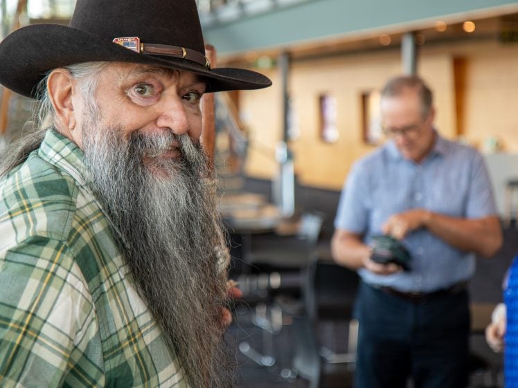Image of An older man with a long grey beard, wearing a checked shirt and a cowboy hat, smiles at the camera. In the blurred background, two people are talking and looking at a mobile in a modern indoor setting.