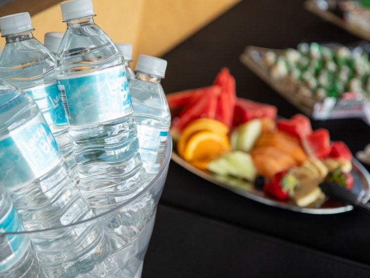Image of A clear ice bucket filled with bottled water sits next to a platter of assorted fresh fruit, including watermelon, oranges, strawberries, and melon slices, on a black tablecloth.