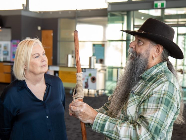 Image of A woman with blonde hair listens intently to a man with a long grey beard and hat who is holding a wooden staff, inside a modern building with large windows.