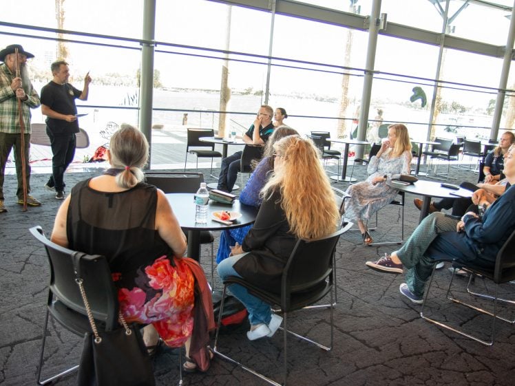 Image of A group of people sit around tables in a bright, modern room with large windows, listening to two presenters—one in a cowboy hat—standing at the front. A scenic view of water and trees is visible outside.