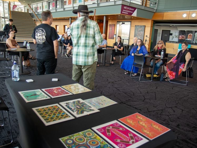 Image of A group of people sit in a semicircle in a large indoor space, listening to two standing presenters. Colourful patterned artworks are displayed on a table in the foreground.