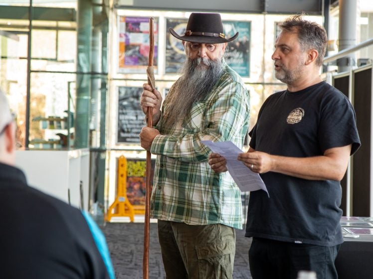 Image of Two men stand indoors; one wears a checked shirt and cowboy hat, holding a wooden staff, while the other, in a black T-shirt, reads from papers. They appear to be addressing a seated group.