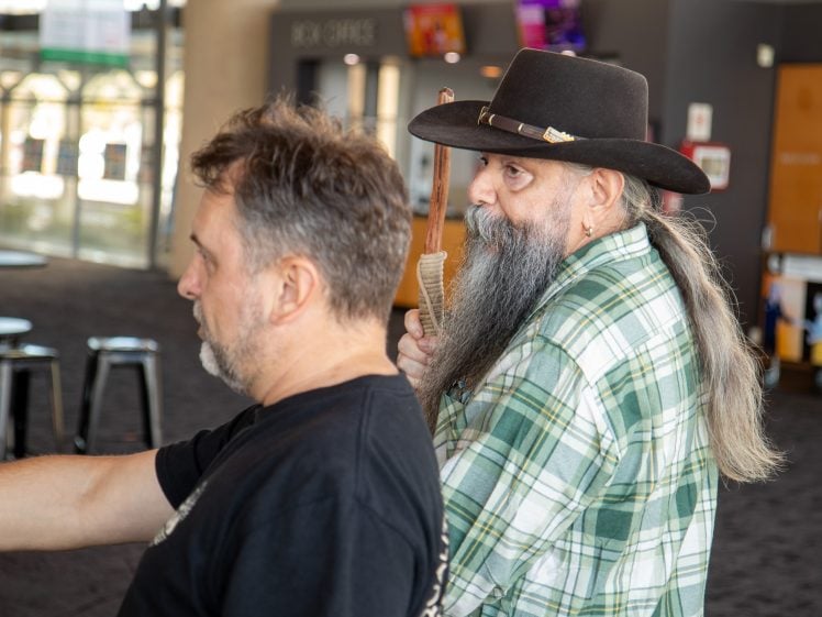 Image of Two men indoors; one in the foreground with short hair wearing a black shirt, the other behind him with long grey hair, a beard, a green checked shirt, and a black hat, holding a wooden stick. The setting appears to be a public space.