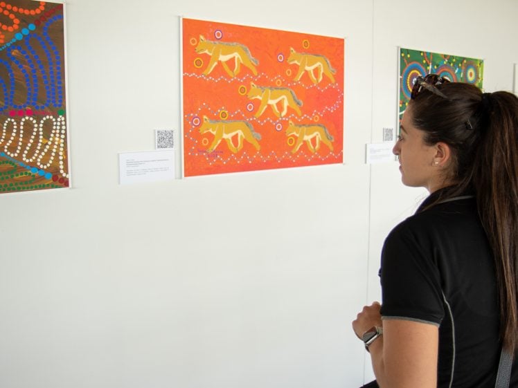 Image of A woman with long brown hair in a ponytail stands in front of a white wall, observing colourful paintings, including an orange artwork with abstract designs and animal shapes.