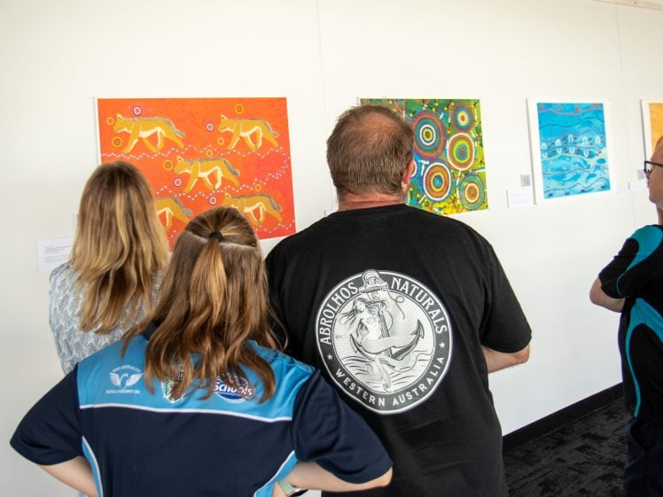 Image of Four people stand in front of a wall, viewing colourful Aboriginal-style artworks displayed in a gallery setting. The viewers' backs are facing the camera, and the room is well-lit.