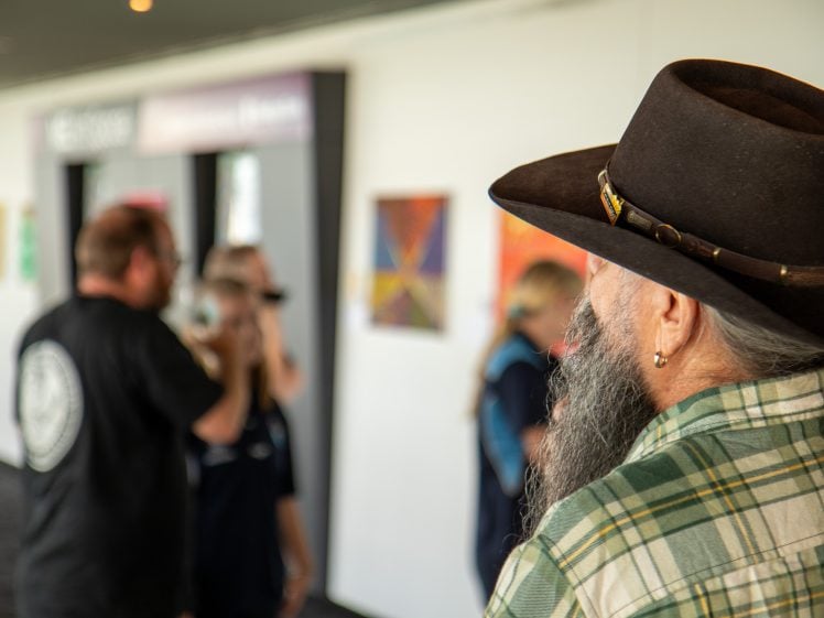 Image of A man with a long grey beard and a brown hat, wearing a green checked shirt, stands in the foreground at an art gallery. In the blurred background, people observe colourful artwork on the walls.