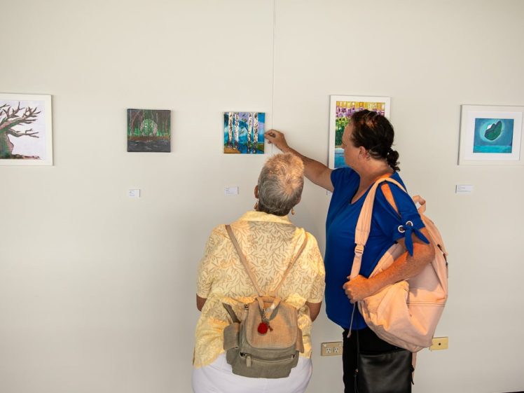 Image of Two people stand in front of a wall displaying small paintings in a gallery. One person points at a painting whilst the other looks on. Both carry rucksacks and face away from the camera.