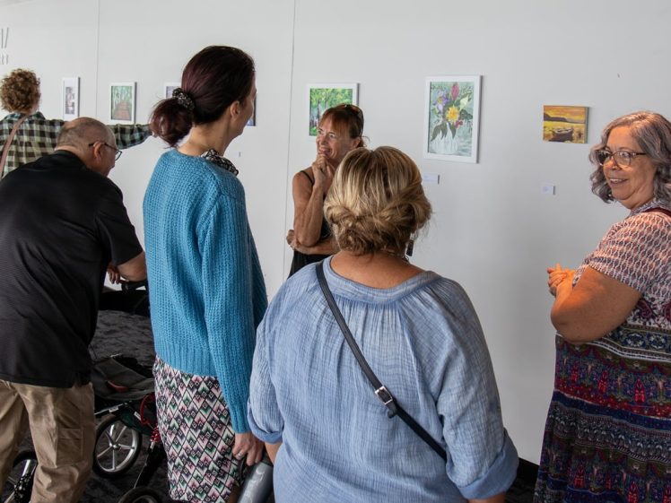 Image of A group of six people stand in front of a white wall displaying small framed artworks, engaging in conversation and viewing the art at an exhibition.