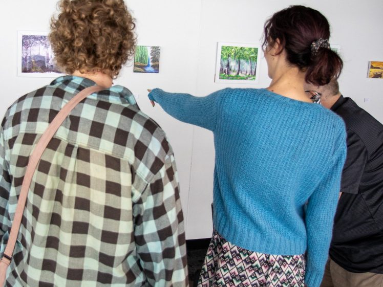 Image of Three people stand in front of a wall displaying colourful landscape photographs; one person in a blue jumper points towards a photo, while the others observe the artwork.