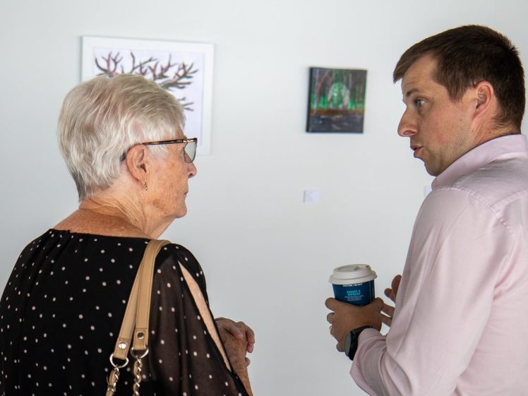 Image of An older woman and a younger man stand facing each other and talking in an art gallery. The man holds a takeaway coffee cup, and paintings are visible on the white wall behind them.