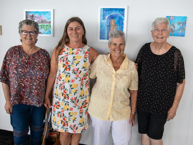 Image of Four smiling women stand side by side in front of a white wall with colourful artwork hanging behind them. They are dressed in casual, summery clothes and appear to be enjoying their time together.