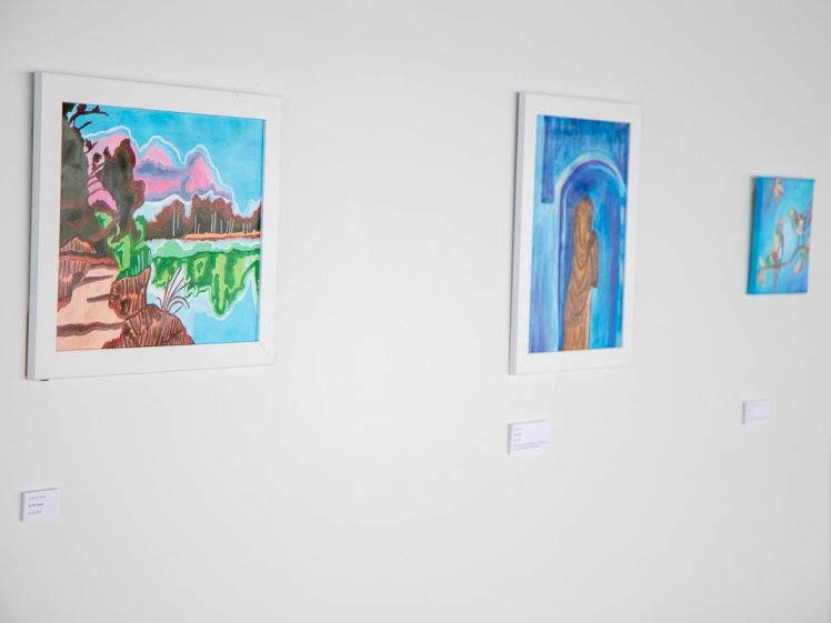 Image of Three colourful framed paintings hang on a white wall in a gallery, each with a small label beneath. The art includes a landscape, an abstract piece with a blue arch, and a small painting featuring blue and pink tones.