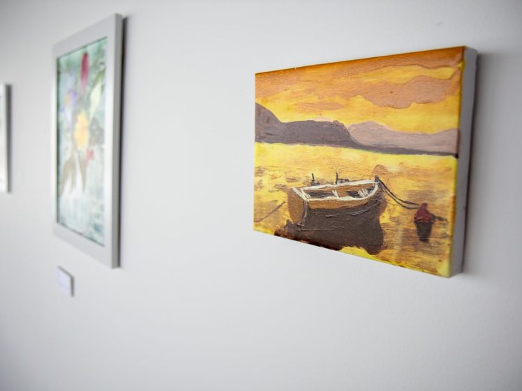Image of A painting of a boat on a lake at sunset hangs on a white wall alongside other framed artworks in a gallery setting.