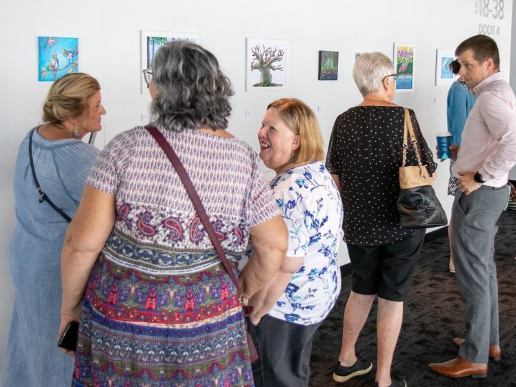 Image of A group of people stand and chat while viewing colourful artworks on a gallery wall. Some are engaged in conversation, while others closely examine the paintings. The scene appears bright and social.